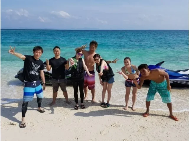 晴天の沖縄のビーチで、水着やラッシュガード姿で笑顔でピースサインをする男女7人のグループ。背景にはエメラルドグリーンの海とジェットスキー。
