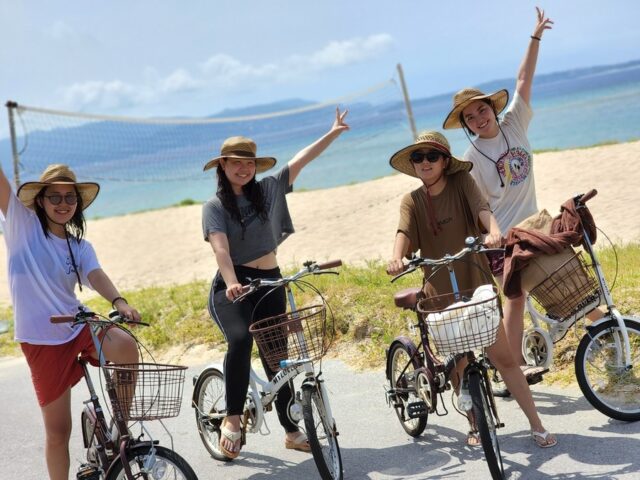 伊江島の白い砂浜を背景に、レンタサイクルを楽しむ笑顔の女性グループ