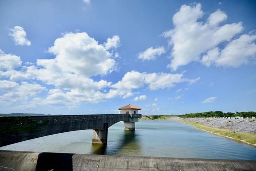 青空と白い雲の下、穏やかな水面に浮かぶ赤瓦屋根の取水塔。沖縄ののどかなダム湖の風景。