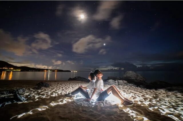 恩納村の夜のビーチで月明かりの下背中合わせに座るカップル