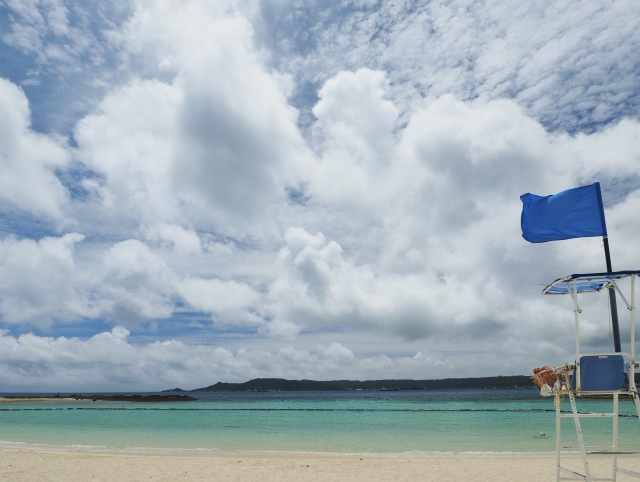 青い空と白い砂浜が広がる沖縄のビーチ。監視台に掲げられた青い旗と穏やかなエメラルドグリーンの海