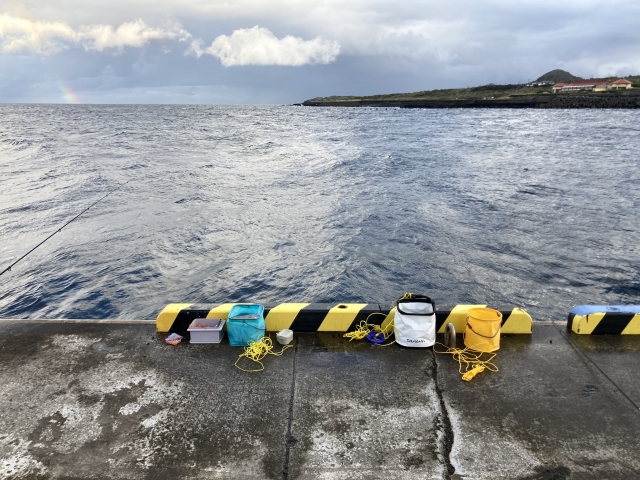 虹の見える沖縄の堤防に並べられた釣り道具一式（バッカンや水汲みバケツ）
