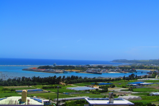 高台から見下ろす沖縄の漁村と海。サンゴ礁の広がる浅瀬の海と、緑豊かな農地が混じり合う美しいパノラマ。