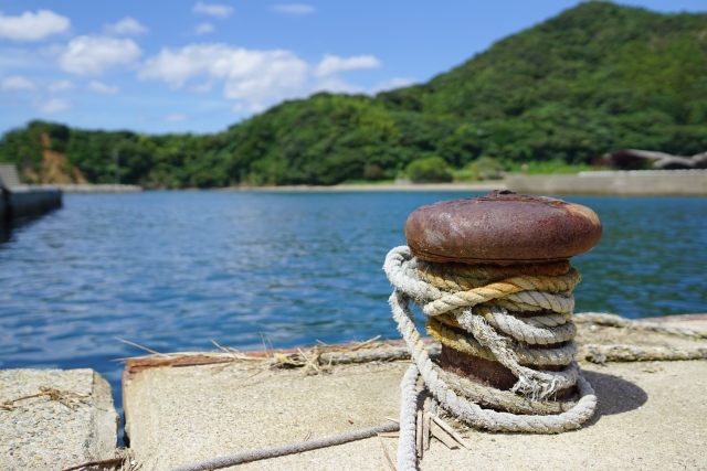 漁港の岸壁にある、ロープが何重にも巻き付けられた古い係留用ボラードのアップ。背景に広がる海と山。