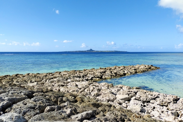 岩場の海岸から望む、エメラルドグリーンの海と水平線に浮かぶ伊江島（城山）
