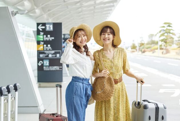 空港のターミナル前で、キャリーバッグを手に笑顔で並ぶ麦わら帽子の女性二人組。沖縄旅行の始まり。